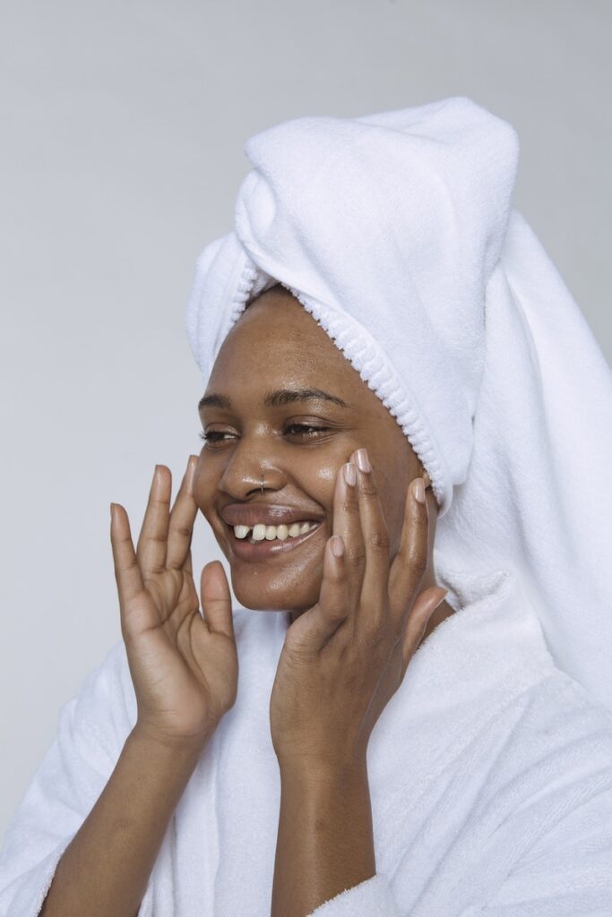 Side view of smiling black female in white bathrobe and towel on head smearing cream on face and looking away against white background